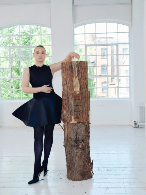 A young woman in a black dress and dark tights leans on stacked logs in a white photo studio with large windows. Fashion editorial.