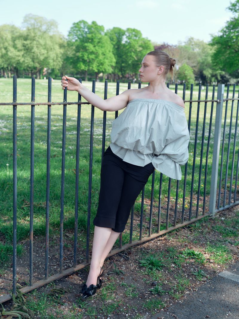 Blonde woman in off-shoulder top and dark pants leaning against a rusty metal fence in an outdoor park-like environment with green trees.