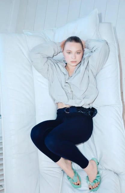 Overhead shot of a woman lying on a white couch, wearing a gray cropped jacket, dark leggings, and light blue sandals. Hands behind head.
