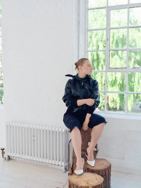 Stylish woman in black jacket and dark culottes poses seated on a pair of log stools in a bright interior space near a window.