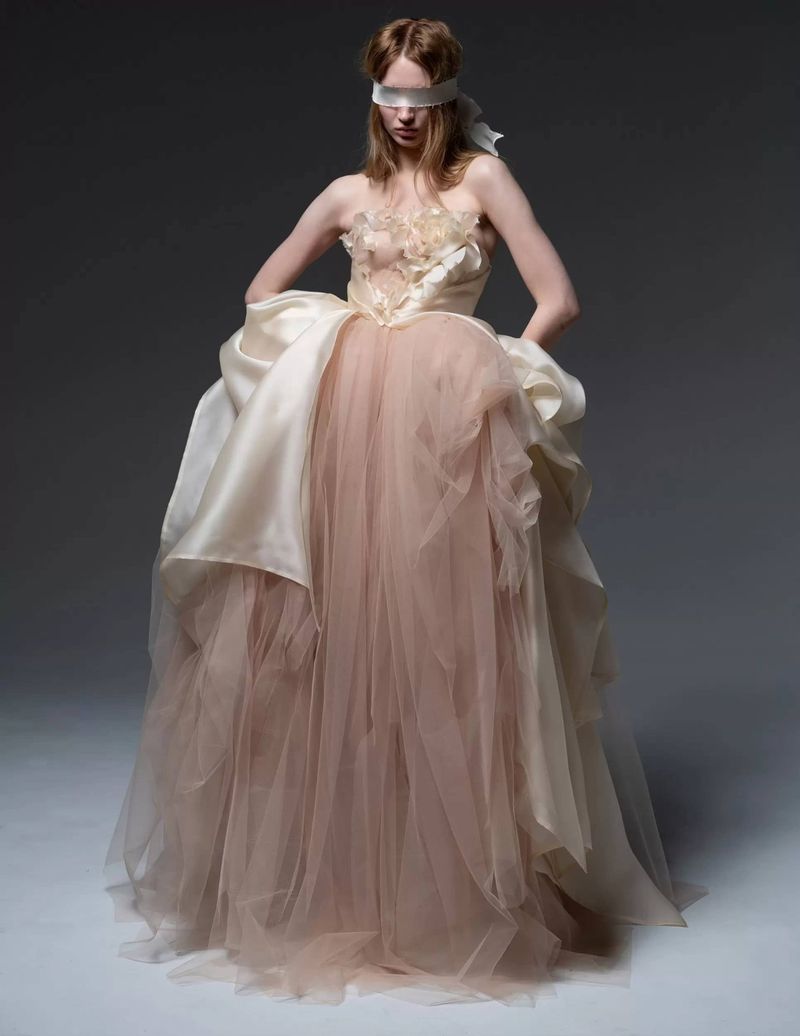 Woman in a strapless tulle gown with a draped bodice and a satin blindfold poses in a studio against a neutral gray background.