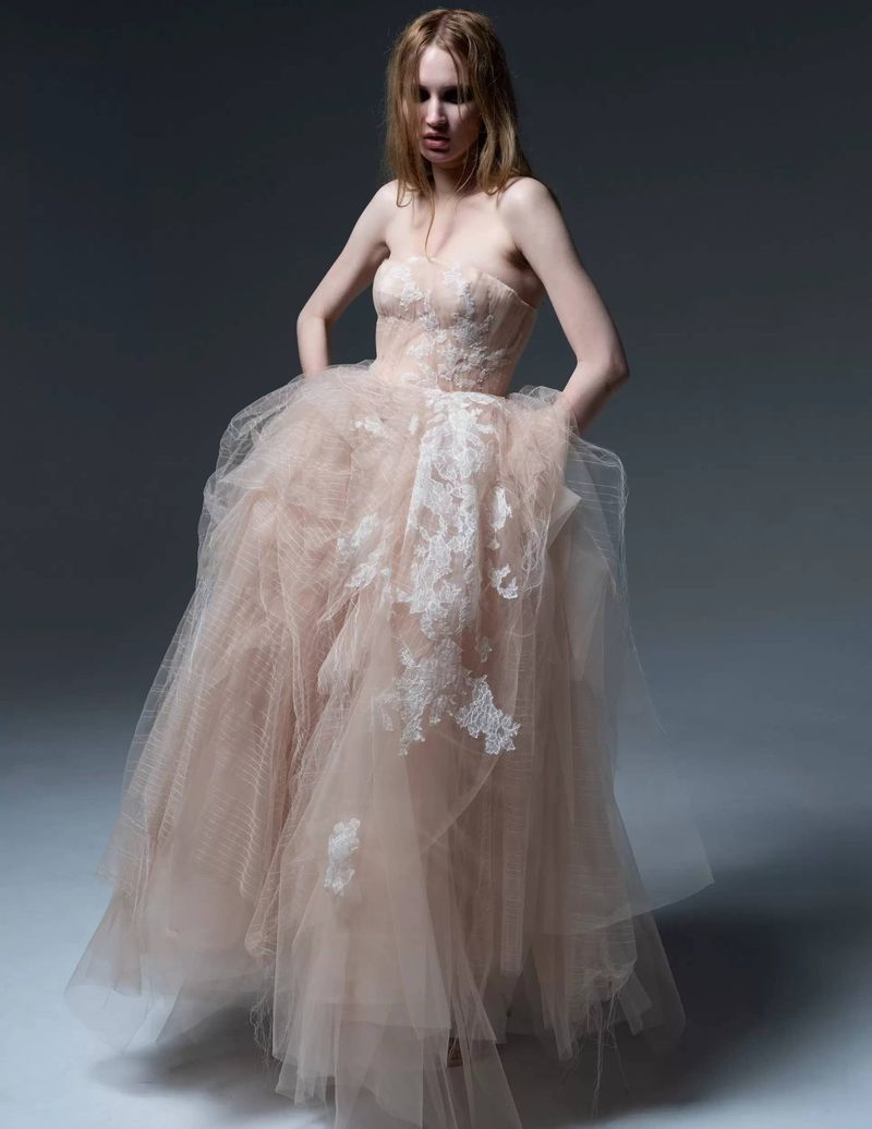Adult woman in a strapless pale-pink tulle gown with lace appliqués posing in a photo studio with a gray backdrop.
