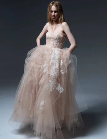 Adult woman in a strapless pale-pink tulle gown with lace appliqués posing in a photo studio with a gray backdrop.