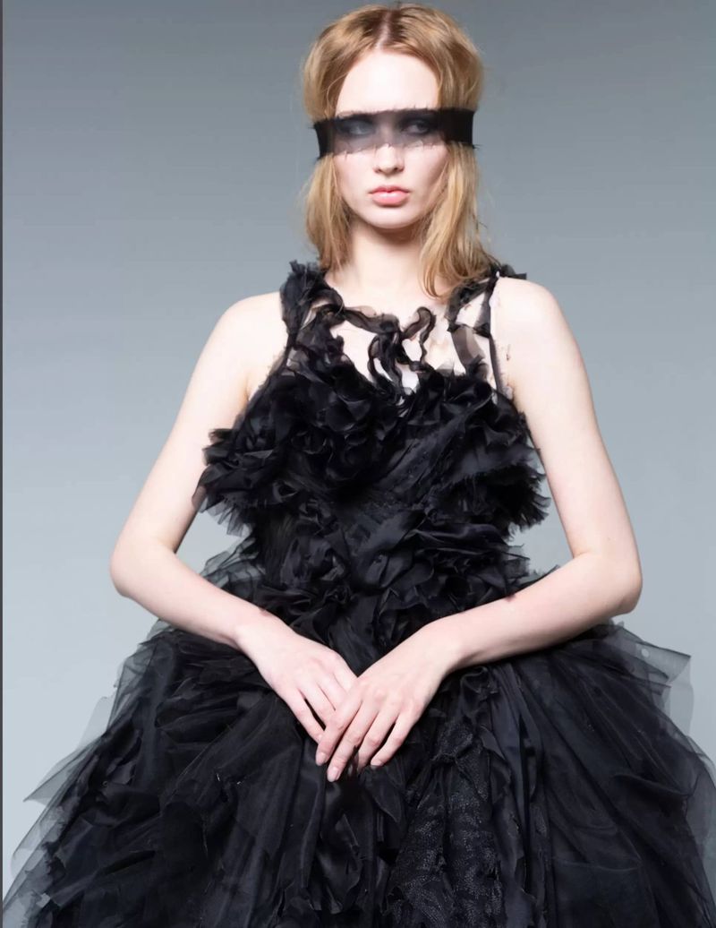 Woman wearing a black tulle dress with a matching blindfold poses in front of a grey backdrop, creating a dramatic fashion portrait.