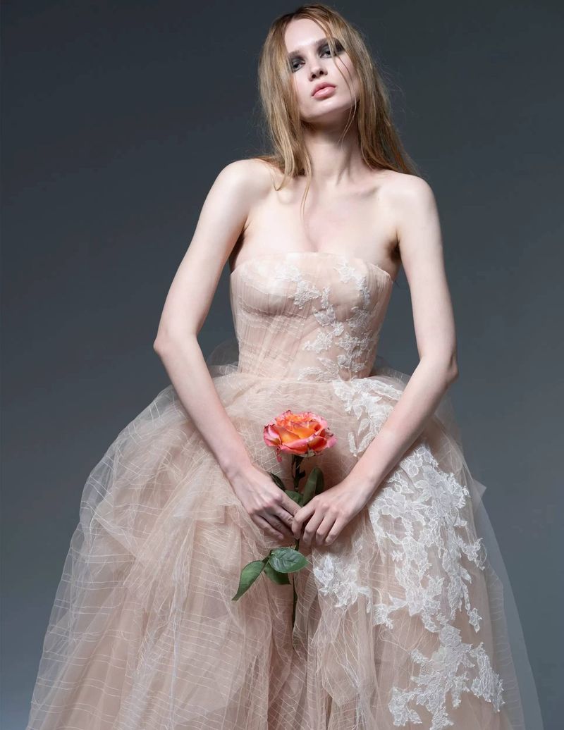 Woman in a strapless peach tulle gown with lace, holding an orange rose with green leaves in a grey studio setting.