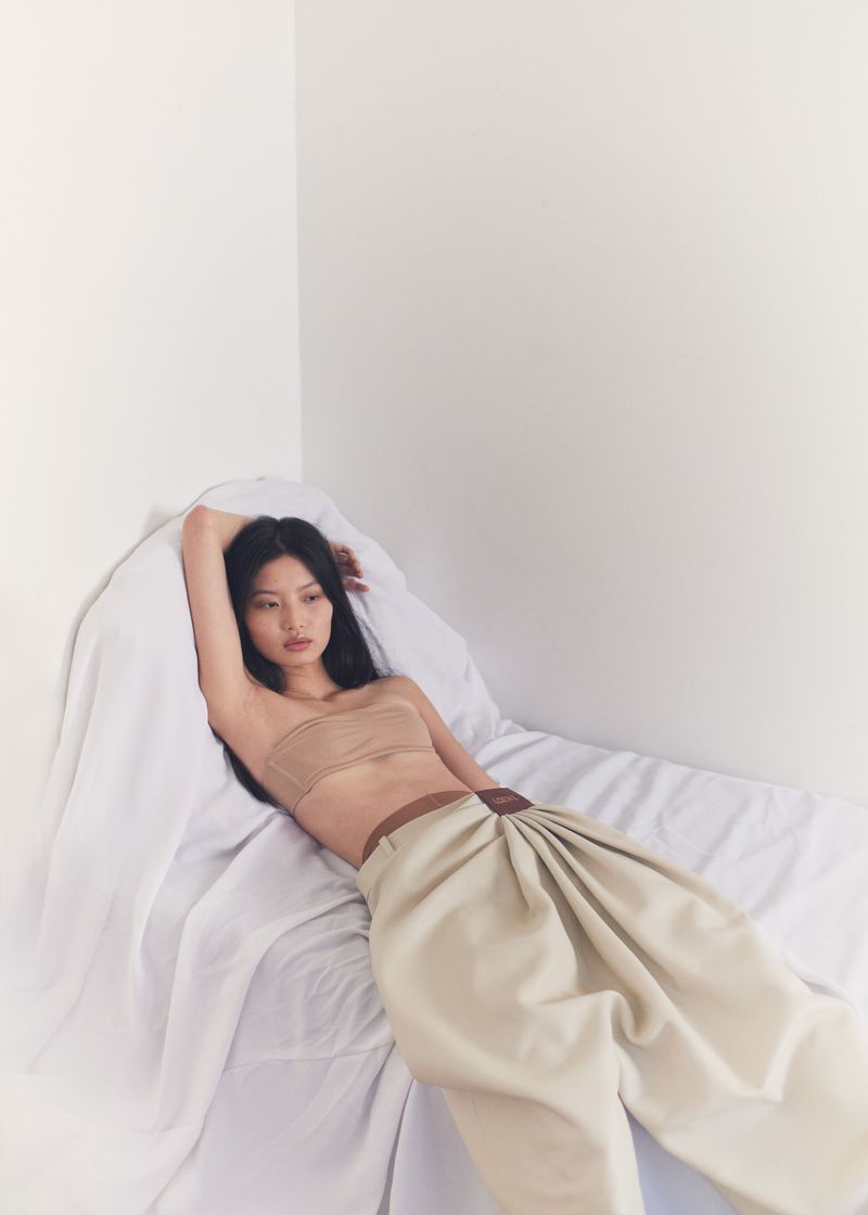An Asian woman reclines on a white sheet in a photo studio, wearing a tan bandeau top and high-waisted, pale beige pants.