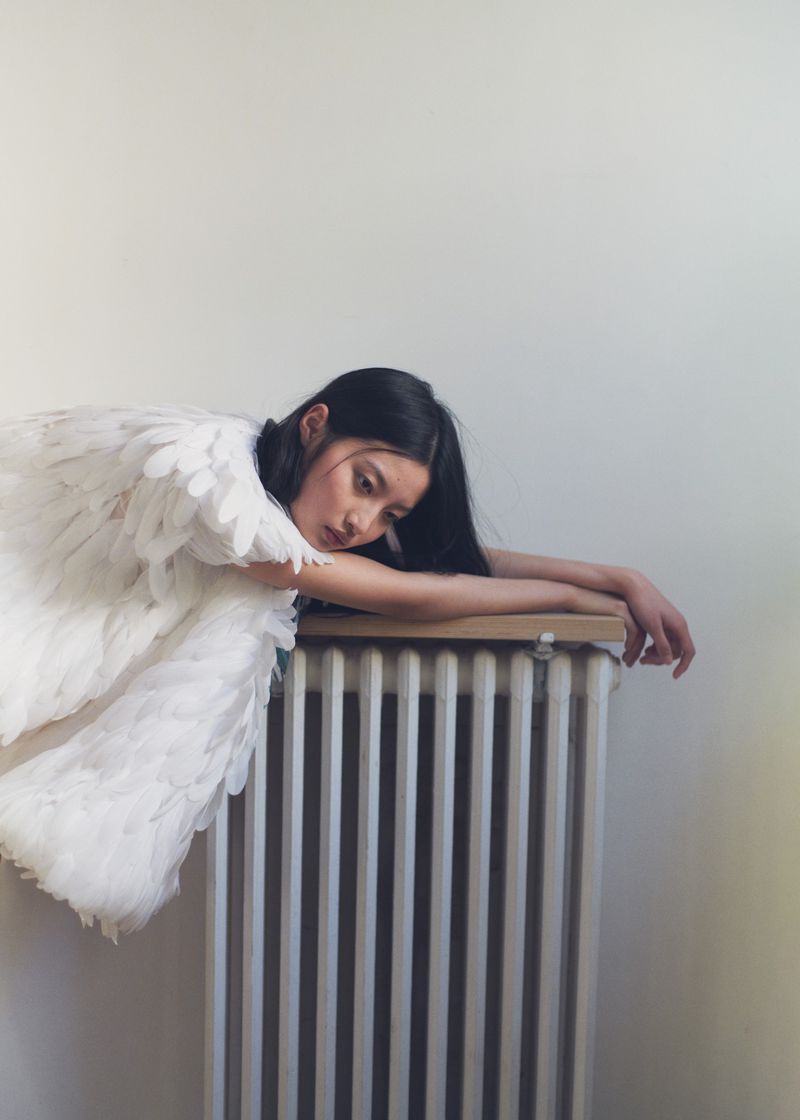 An Asian woman with long black hair wearing white feathered angel wings rests her arms and head on a vintage white radiator.