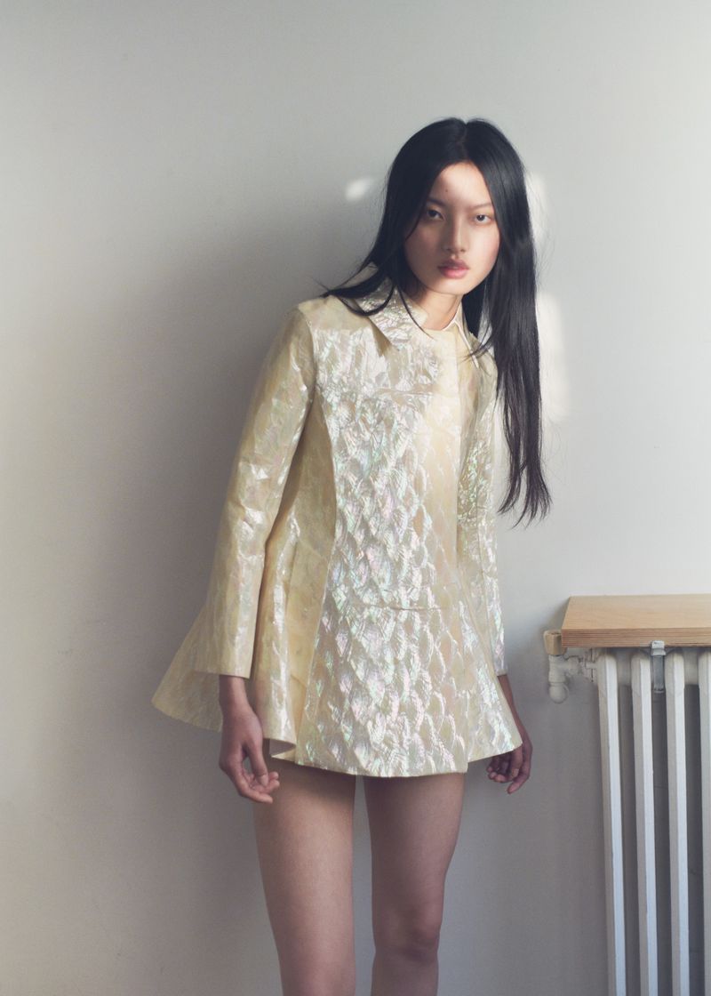 An Asian woman with long black hair poses in a shimmering, cream-colored, short dress against a white wall, near a white radiator.