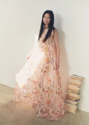 Model Chen Yang in a long floral dress standing beside a stacked pile of books against a white wall.