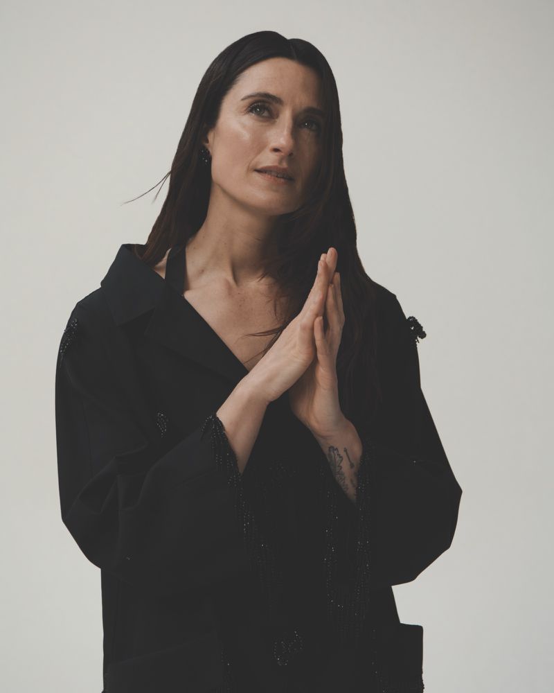 Portrait of a woman with long dark hair wearing a black jacket against a white backdrop in a studio setting with natural lighting.