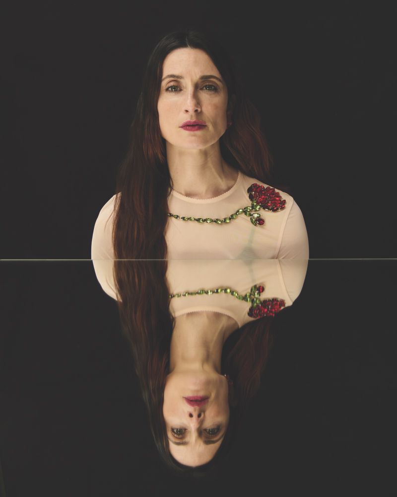 Portrait of a fair-skinned woman with long brown hair in a light pink top with rose embellishments, reflected in a dark mirror.