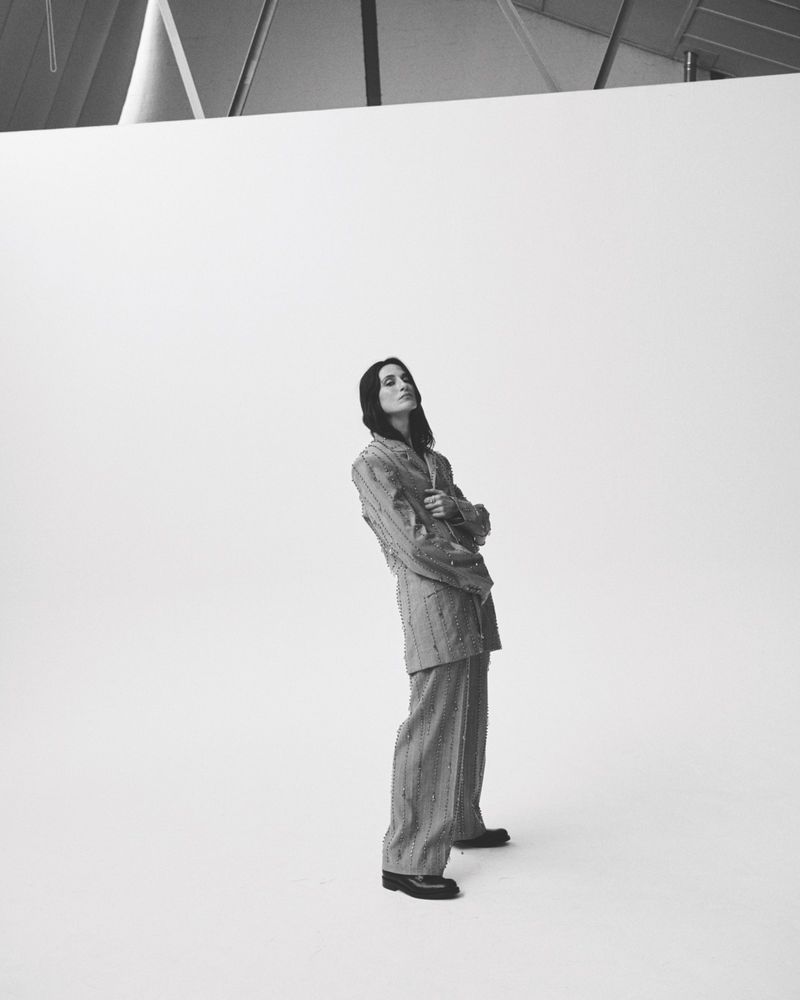 Monochrome portrait of a person standing with arms crossed in a pinstripe suit in front of a white and geometric background.