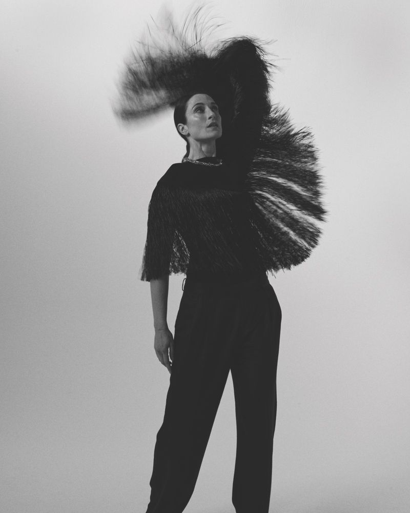 Portrait of a stylish woman in a black outfit with a large feathered headpiece, striking a pose against a simple white background in a studio setting.
