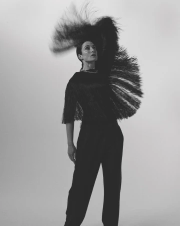 Portrait of a stylish woman in a black outfit with a large feathered headpiece, striking a pose against a simple white background in a studio setting.