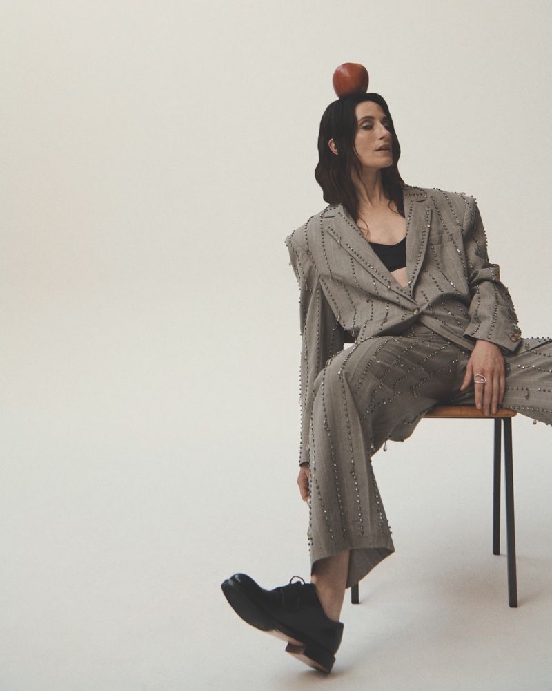 A person in a grey embellished suit sits on a chair with a red apple balanced on their head against a pale backdrop in a studio.