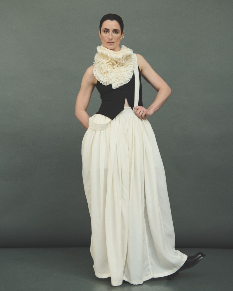 A woman models an avant-garde fashion outfit with a black top, flowing white skirt, and ruffled collar in a neutral studio setting.