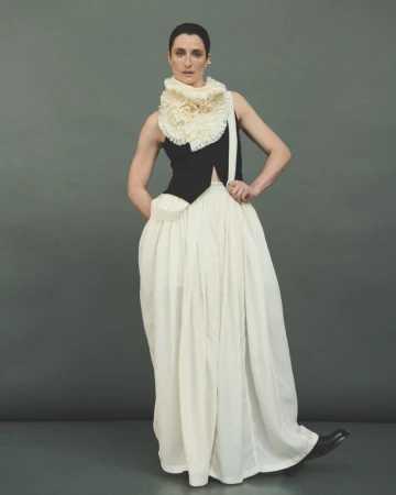 A woman models an avant-garde fashion outfit with a black top, flowing white skirt, and ruffled collar in a neutral studio setting.