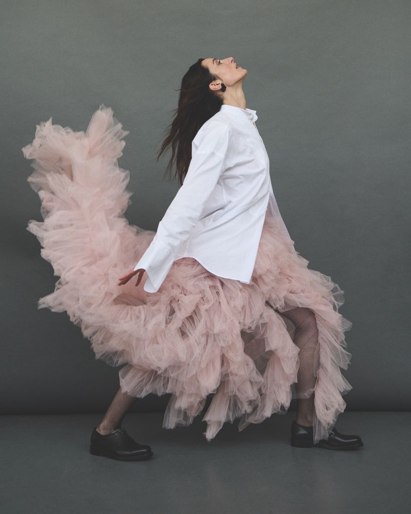 A woman in a white shirt and pink tulle skirt poses in a studio with a gray background. She wears black shoes and has dark hair.