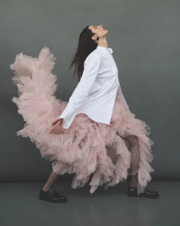 A woman in a white shirt and pink tulle skirt poses in a studio with a gray background. She wears black shoes and has dark hair.