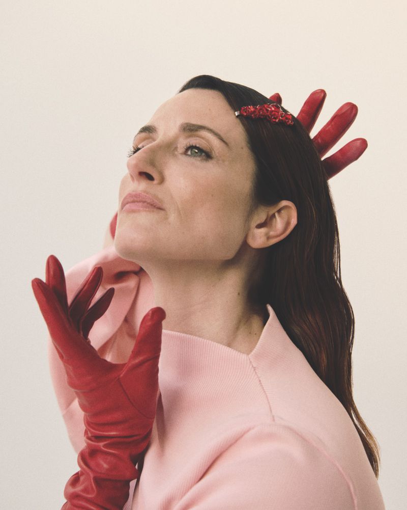 A fashionable woman with dark hair is looking up. She is wearing a pink sweater and red gloves, styled with a red hair clip.