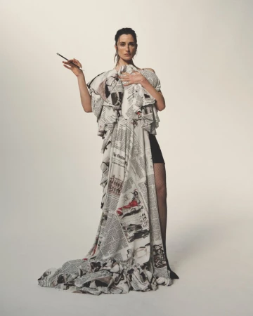A woman poses in a striking dress crafted from newspaper print, accessorized with a cigarette holder, against a stark white background.