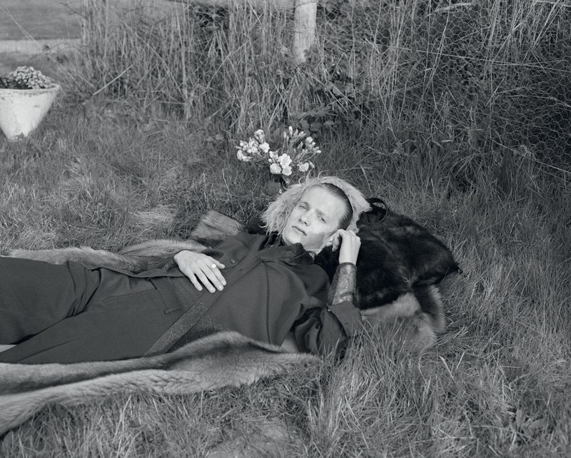 Monochrome portrait of a young adult lying in the grass, wearing a dark jumpsuit, next to a fur throw, with a small flower arrangement.