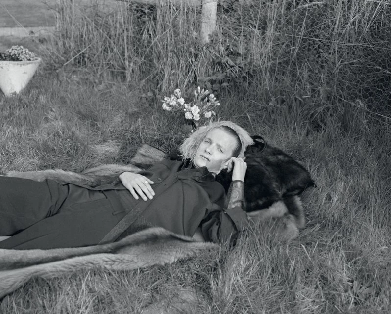 Monochrome portrait of a young adult lying in the grass, wearing a dark jumpsuit, next to a fur throw, with a small flower arrangement.