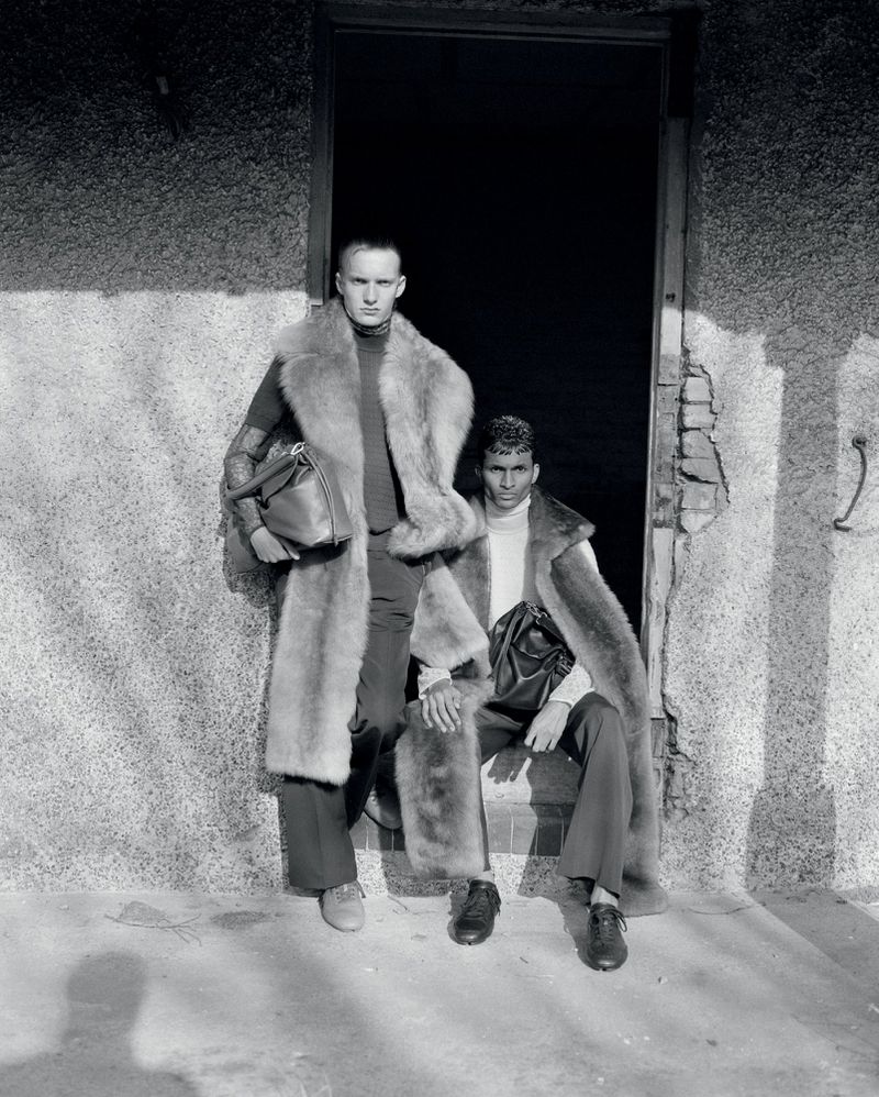 Two adult men in fur coats posing in a doorway with bags, in a black and white fashion editorial style photograph.