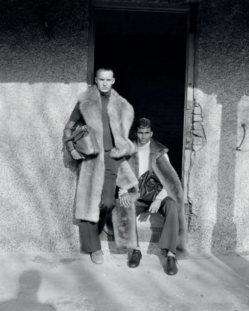 Two adult men in fur coats posing in a doorway with bags, in a black and white fashion editorial style photograph.