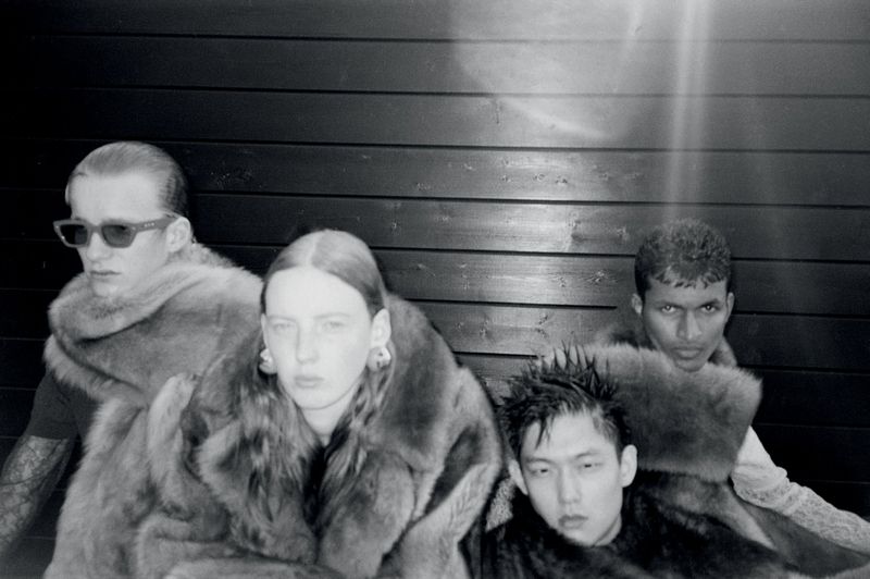 Black and white studio portrait of four diverse fashion models wearing large fur coats against a dark wooden wall background.