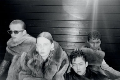 Four models wearing plush fur scarves posing against dark wooden slat walls for Arena Homme + photographed by Paul Wetherell