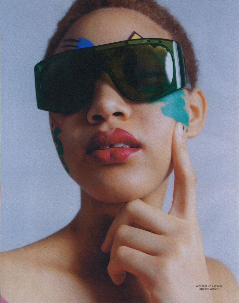 Young woman with creative makeup and oversized sunglasses, posing indoors, creating a fashionable and artistic portrait.