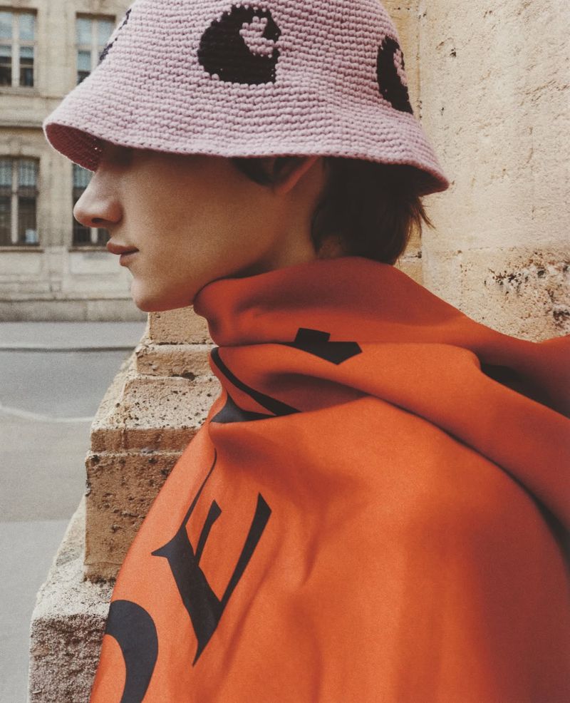 Young adult male in an orange hoodie and pink patterned bucket hat stands next to a textured wall in an outdoor urban environment.