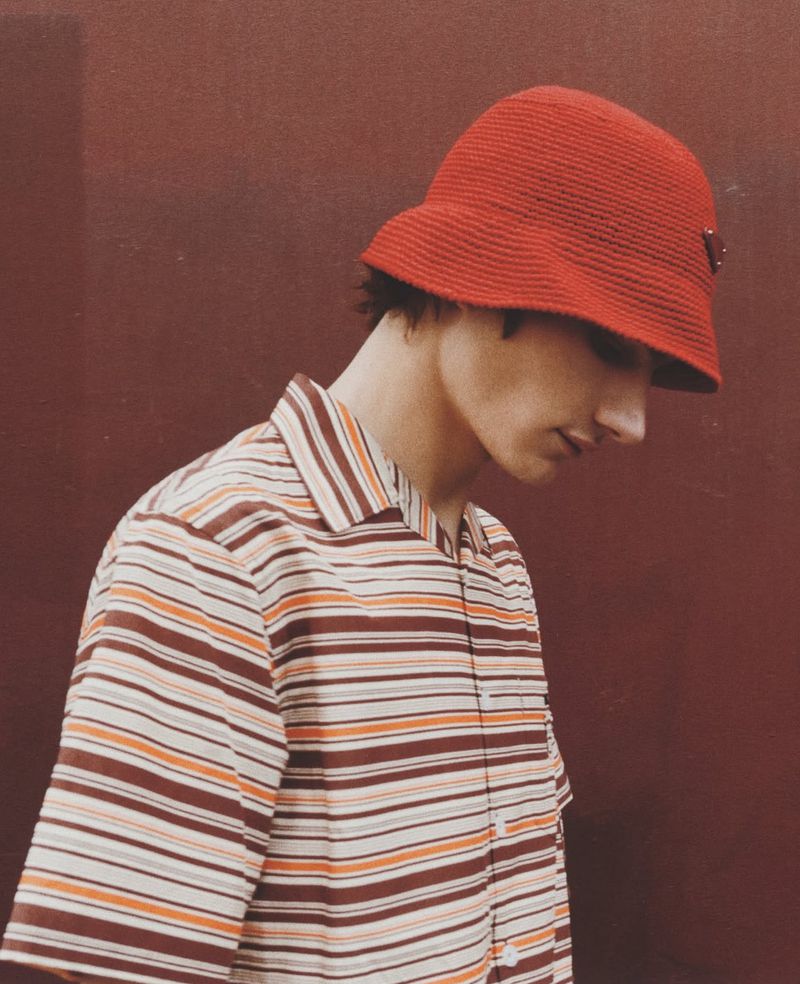 Stylish person poses in red hat and striped shirt against red background indoors. Portrait shot emphasizing fashion details and color palette.