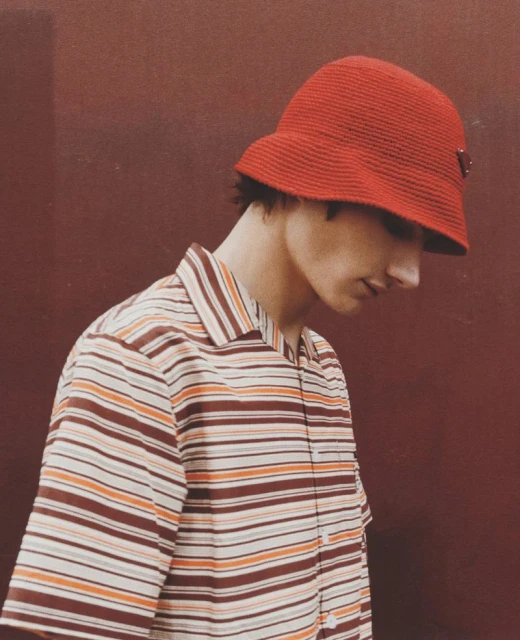 Stylish person poses in red hat and striped shirt against red background indoors. Portrait shot emphasizing fashion details and color palette.