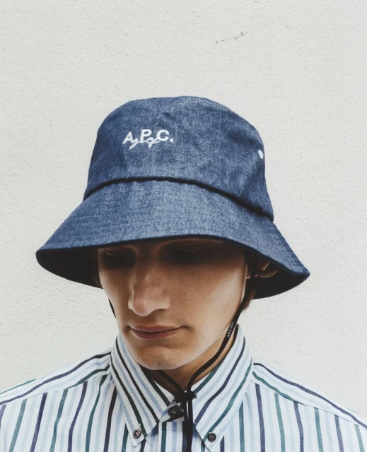 Man wearing blue denim bucket hat and striped button-down shirt in a close-up studio shot against a neutral background.