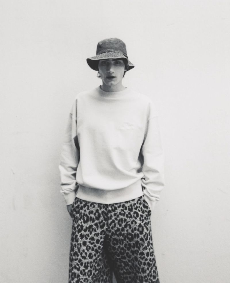Monochromatic portrait of a person wearing a bucket hat, sweatshirt, and leopard-print pants standing against a white wall.