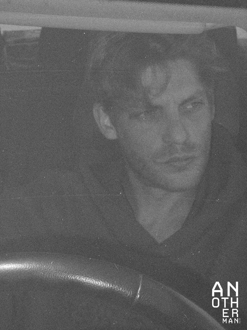 Monochromatic portrait of a man in a vehicle wearing a hooded sweatshirt. The scene is dimly lit, showcasing a steering wheel.
