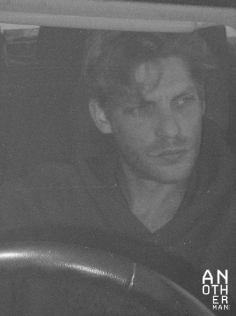 Monochromatic portrait of a man in a vehicle wearing a hooded sweatshirt. The scene is dimly lit, showcasing a steering wheel.