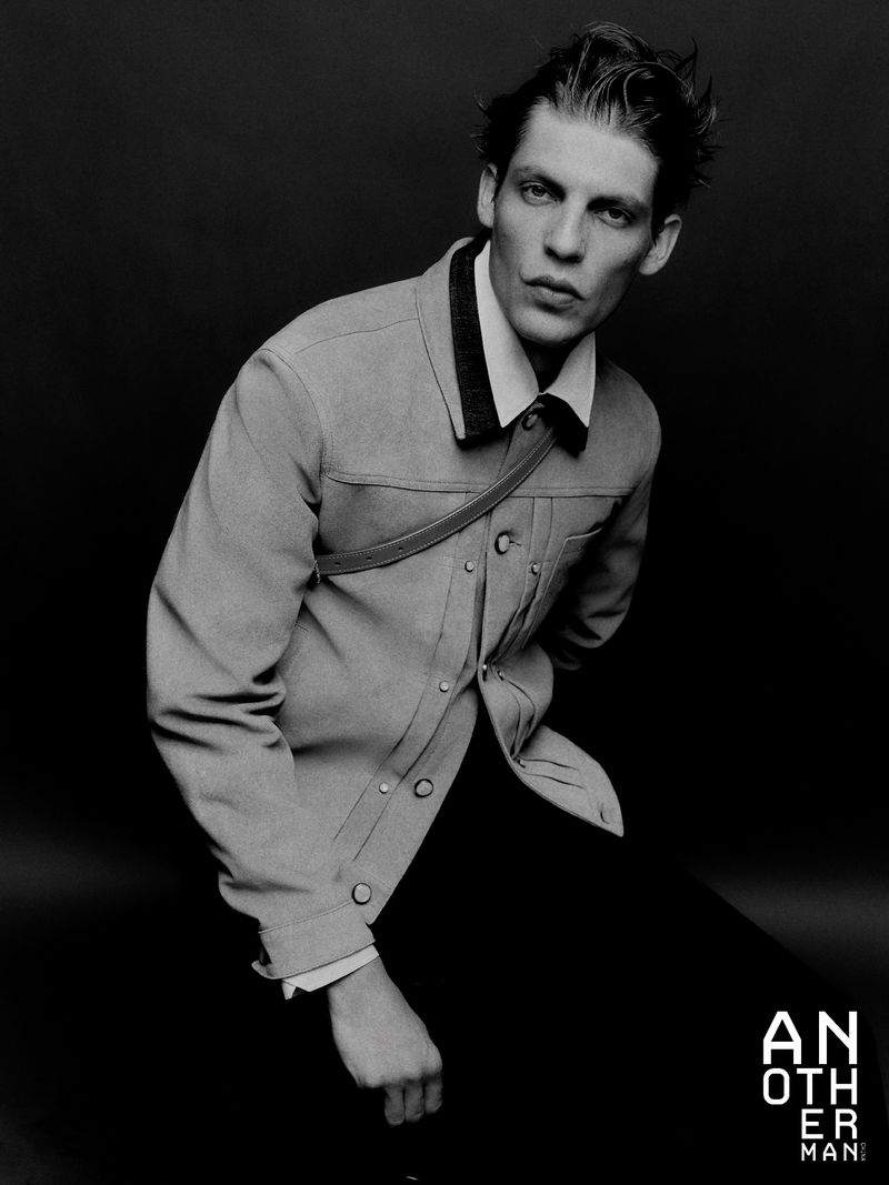 Portrait of a stylish adult male model in a light-colored jacket and dark trousers, posing indoors against a dark background.