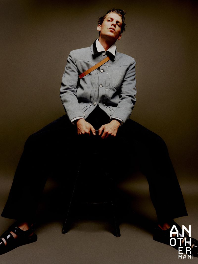 A fashionable young adult male poses while seated, wearing a grey jacket, black trousers, and sandals in a studio setting.