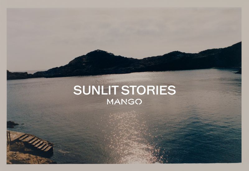 Scenic view of ocean inlet with rocky coastline, calm water reflecting sunlight, and promotional text overlay for Mango's 'Sunlit Stories.'