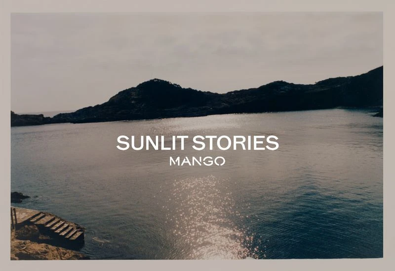 Scenic view of ocean inlet with rocky coastline, calm water reflecting sunlight, and promotional text overlay for Mango's 'Sunlit Stories.'