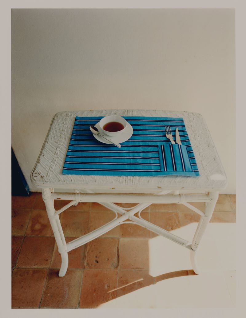 Still life of tea setting on a wicker table. A saucer of tea, silverware and blue striped placemat rest on red brick flooring.