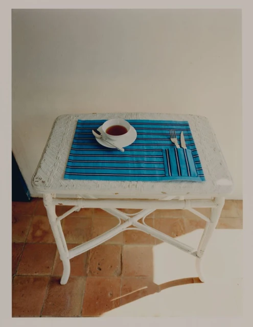 Still life of tea setting on a wicker table. A saucer of tea, silverware and blue striped placemat rest on red brick flooring.
