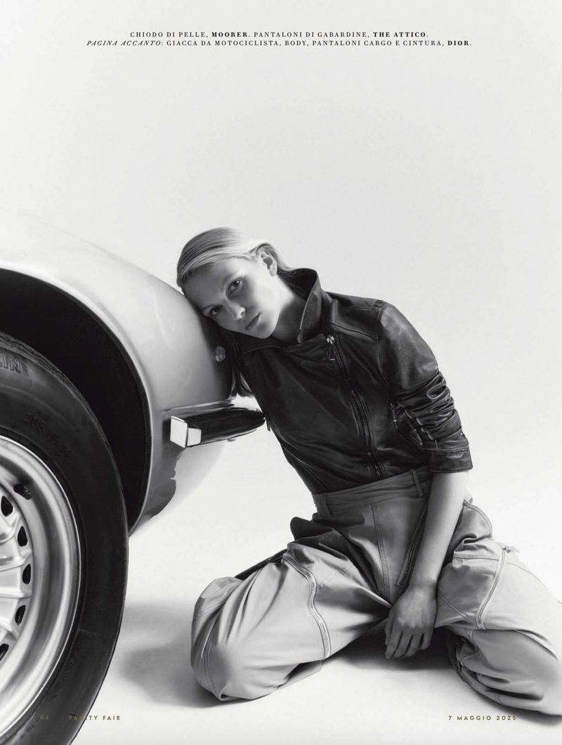 Fashion shot featuring a young adult female model leaning against a vintage car in a minimalist photo studio setting, wearing a dark jacket.