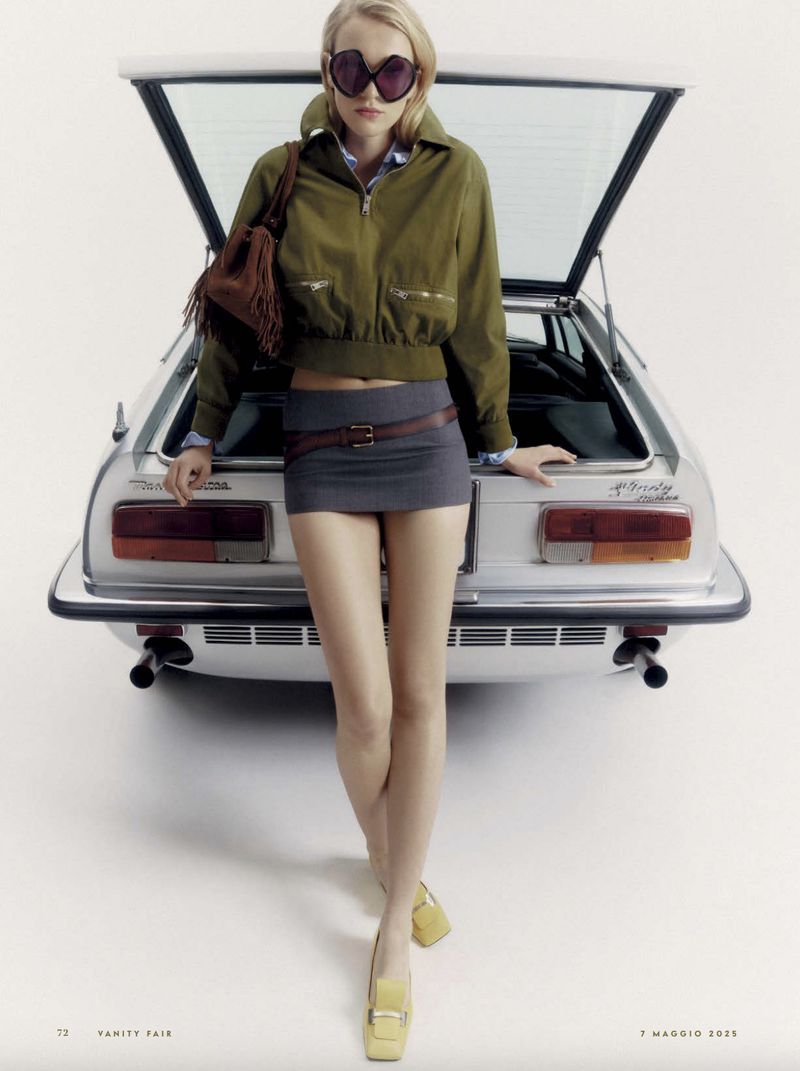 Woman in trendy sunglasses, mini skirt, and jacket poses with a silver sports car against a white backdrop, fashion editorial.