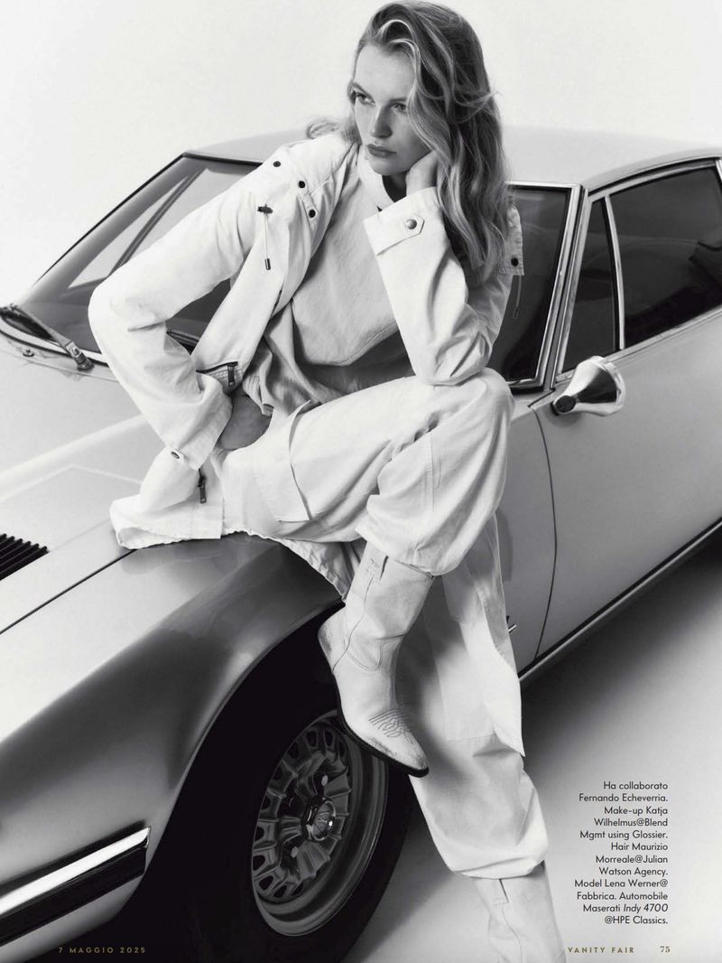 A woman poses on a silver car wearing a white jacket, shirt, and pants, set against a bright studio backdrop in a fashion editorial shot.