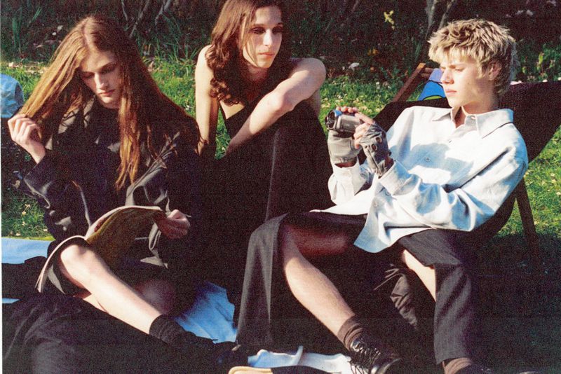 Three young adults are seated on grass, bathed in sunlight. One holds a camcorder, another reads, and the third looks on, wearing black clothing.