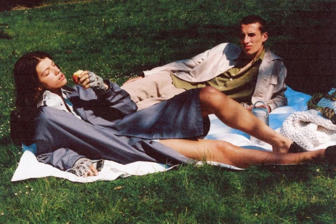 Two people relax on a picnic blanket in a grassy field. One person eats an apple, while the other looks away, enjoying a sunny day.